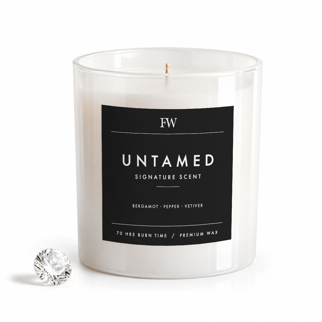A white wax candle in a clear glass jar with a black label reading 'FW UNTAMED SIGNATURE SCENT', shown on a white background with a small faceted crystal-like accent at the lower left.